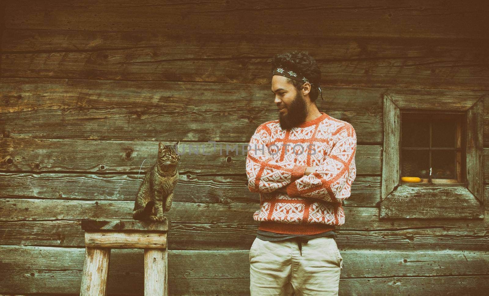 portrait of young hipster man with beard and cat in front of wooden house