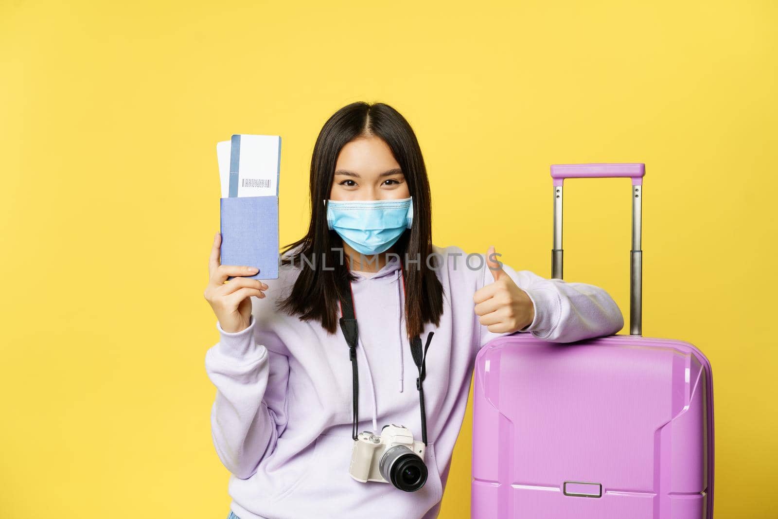 Japanese girl in medical mask, showing her passport and flight travel pass tickets, thumbs up, posing with suitcase, going on vacation, yellow background.