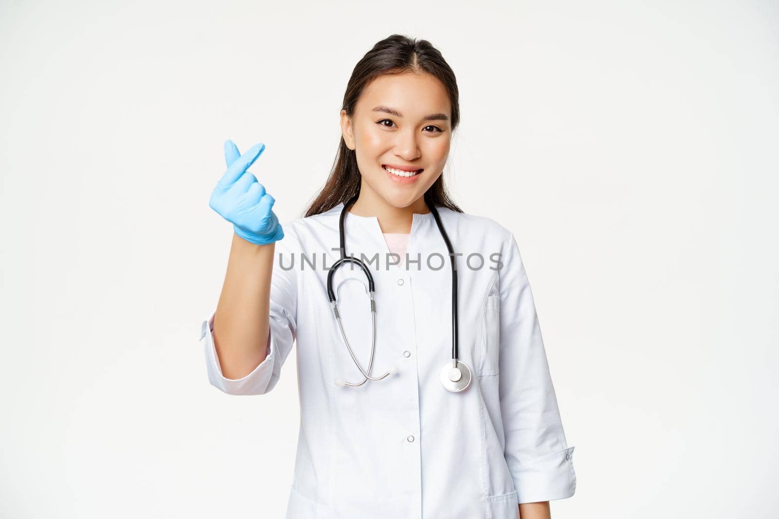 Smiling korean female doctor shows finger heart gesture in rubber gloves, wearing medical uniform, looking happy at camera, white background.