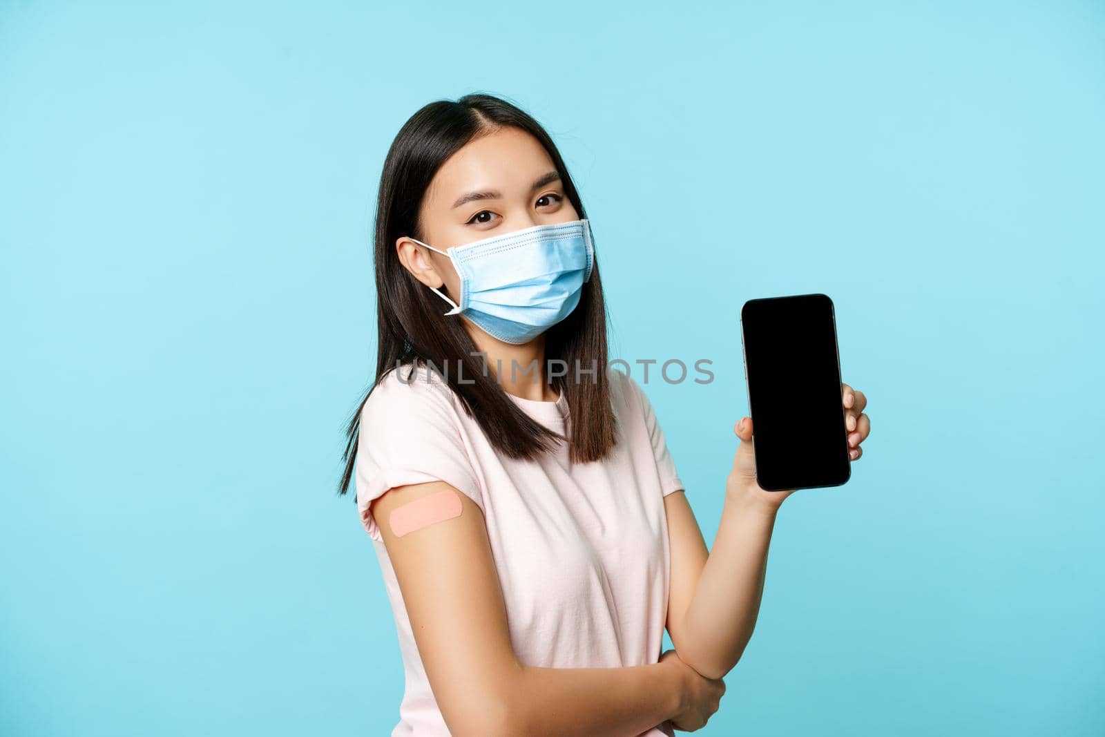 Asian woman in medical face mask, showing arm with patch after covid-19 vaccine, smartphone screen with app interface, concept of vaccination certificate, health passport.