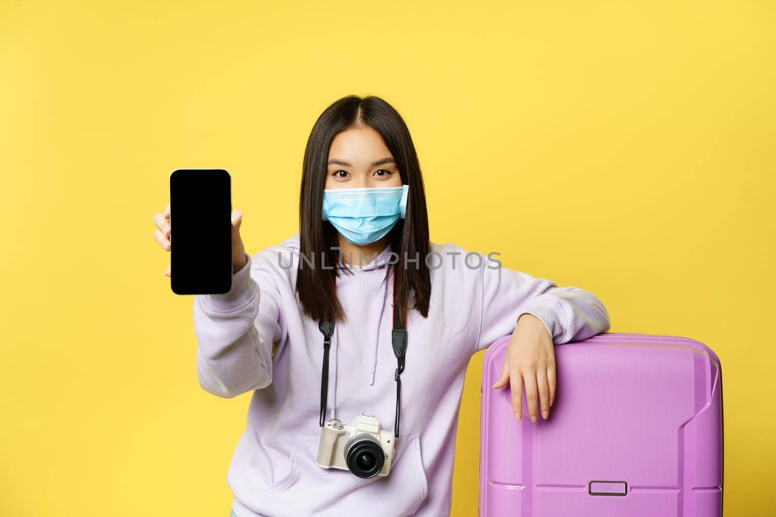 Image of girl traveller, asian tourist shows her phone screen, covid health passport on smartphone app, wearing medical mask and suitcase, yellow background.