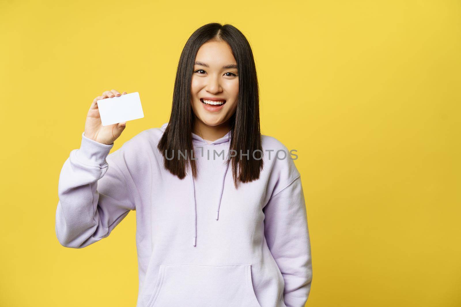 Shopping. Smiling asian woman showing card in her hand, standing over yellow background.