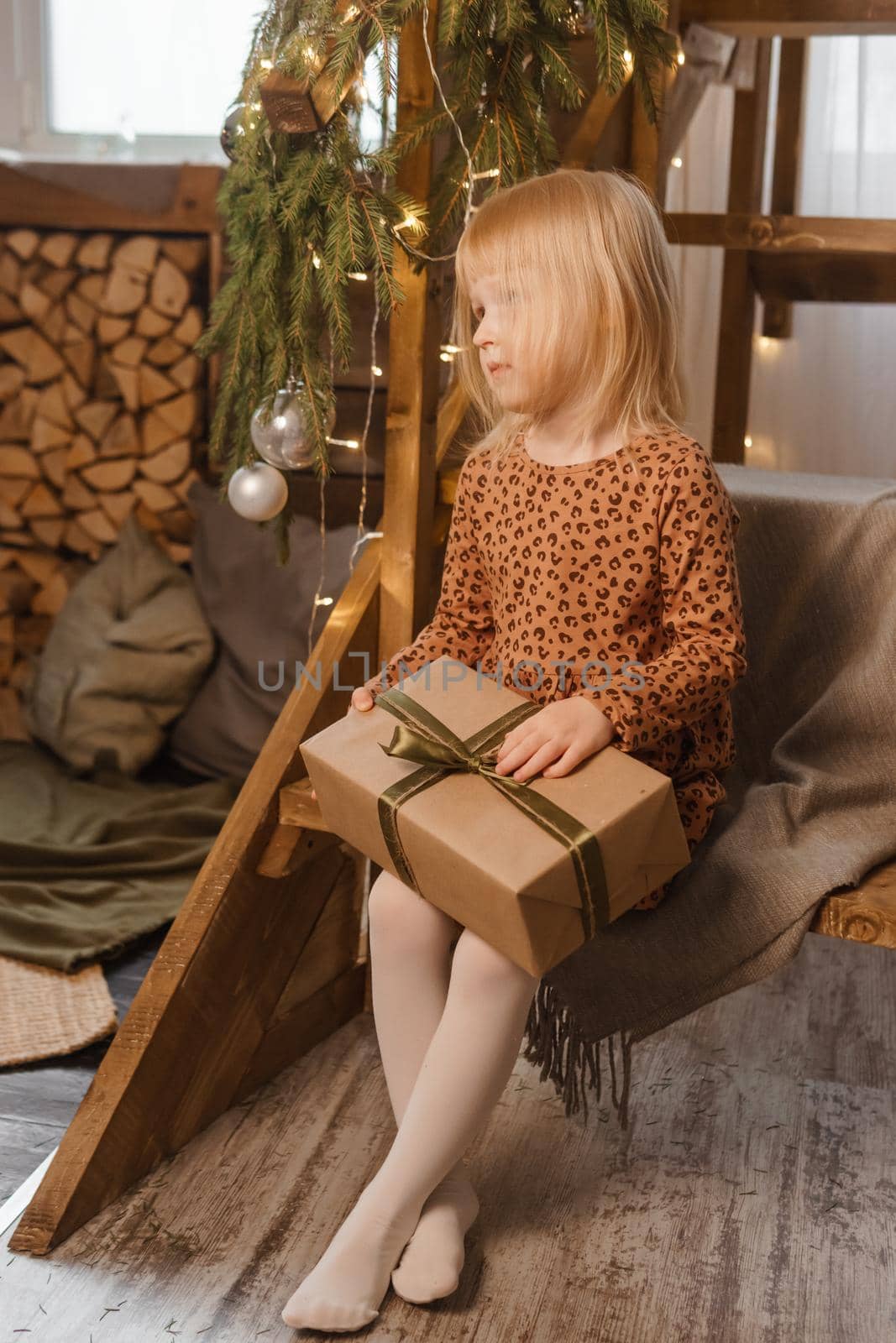 A little blonde girl is sitting on a wooden staircase in a Scandinavian interior decorated in a New Year's style. A child holds a Christmas gift in a craft package.