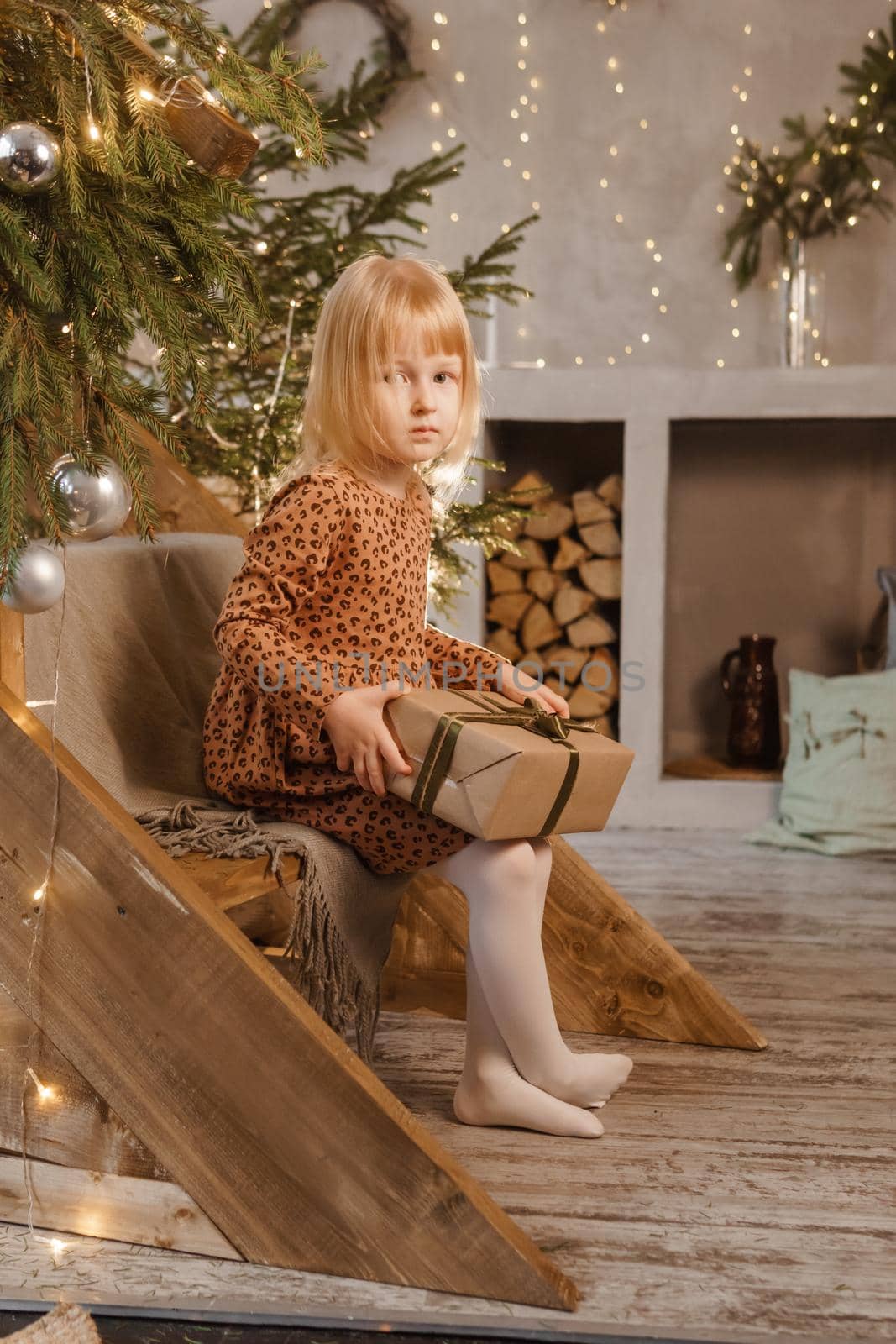 A little blonde girl is sitting on a wooden staircase in a Scandinavian interior decorated in a New Year's style. A child holds a Christmas gift in a craft package.