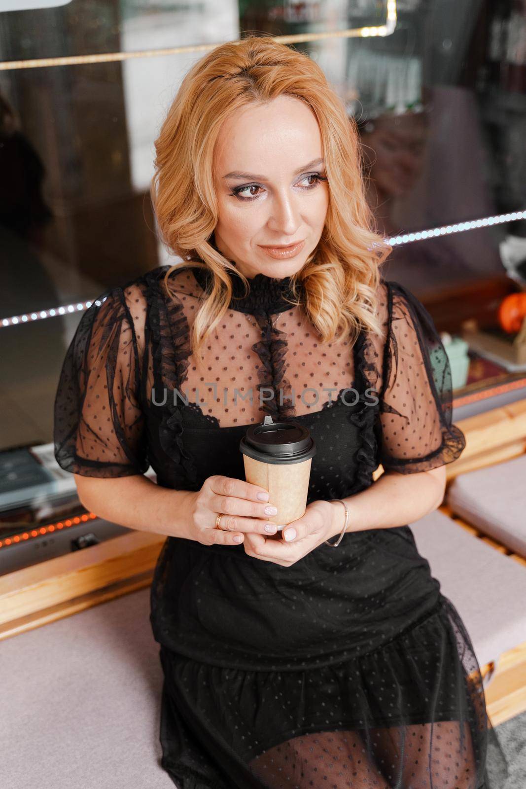 Young charming blonde with a cute smile and makeup while relaxing in a cafe. She is holding a cup of coffee in her hands. She is dressed in a black dress with transparent sleeves