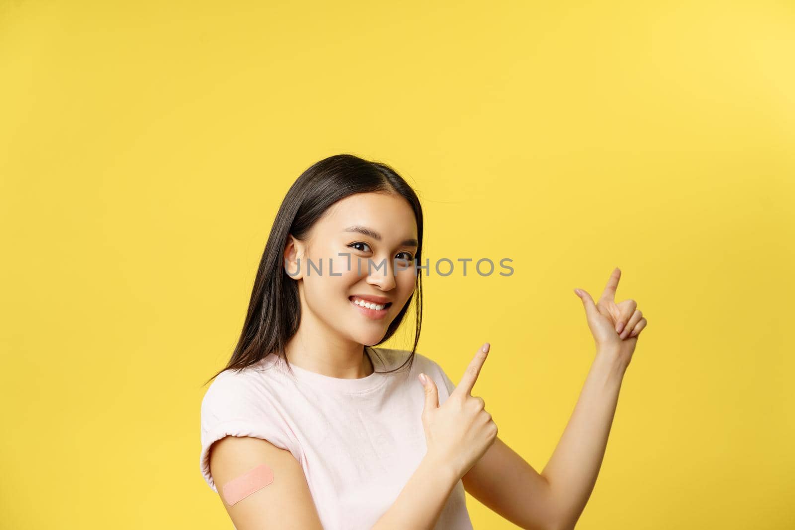 Covid-19 vaccination concept. Cute vaccinated asian woman demonstrating banner, pointing fingers at empty space for promotional text, yellow background by Benzoix