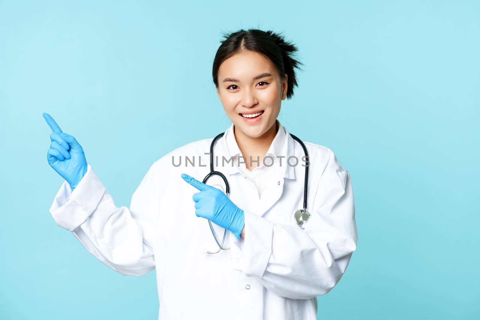 Smiling happy female doctor or nurse, pointing fingers left, wearing medical uniform and gloves, showing hospital advertisement, blue background.