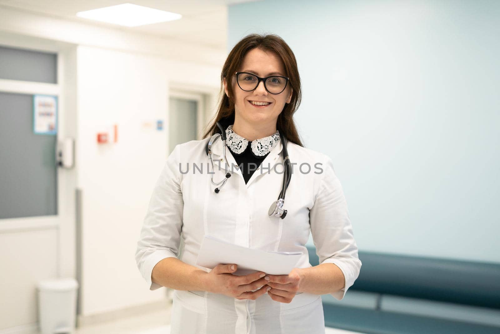 Confident smiling young woman doctor standing in medical institution. Proud professional doctor therapist woman in glasses looking at camera in hospital corridor. Portrait of general practitioner by Tomashevska