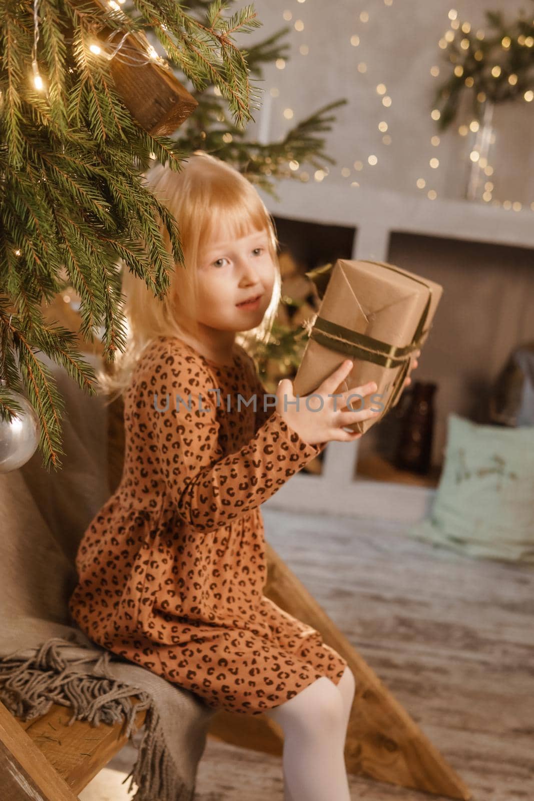A little blonde girl is sitting on a wooden staircase in a Scandinavian interior decorated in a New Year's style. A child holds a Christmas gift in a craft package.