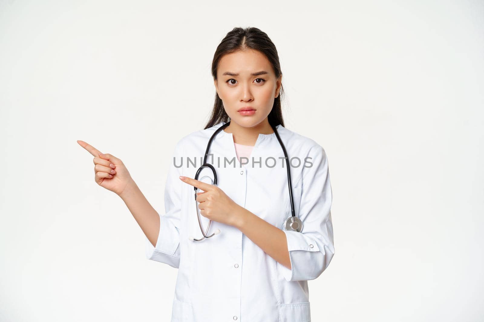 Sad and concerned female doctor, pointing fingers left, showing smth important, promo information, standing over white background.