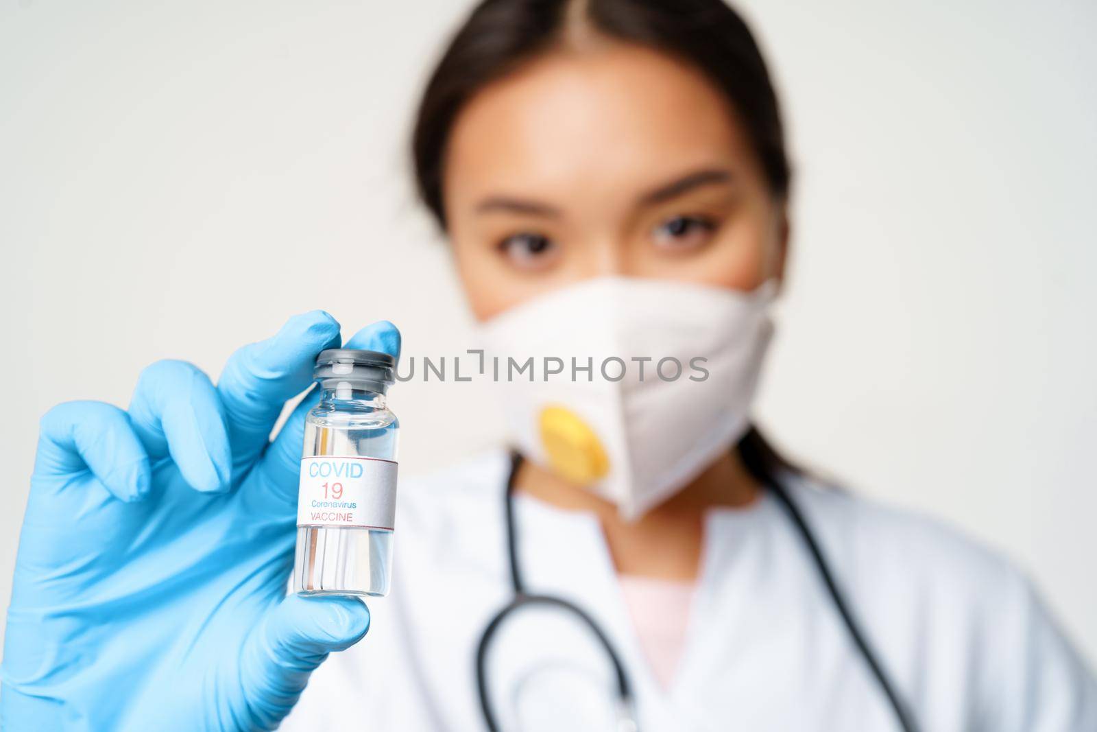 Vaccination and covid-19 prevention concept. Close up portrait of asian doctor in respirator shows bottle with coronavirus vaccine, standing over white background.