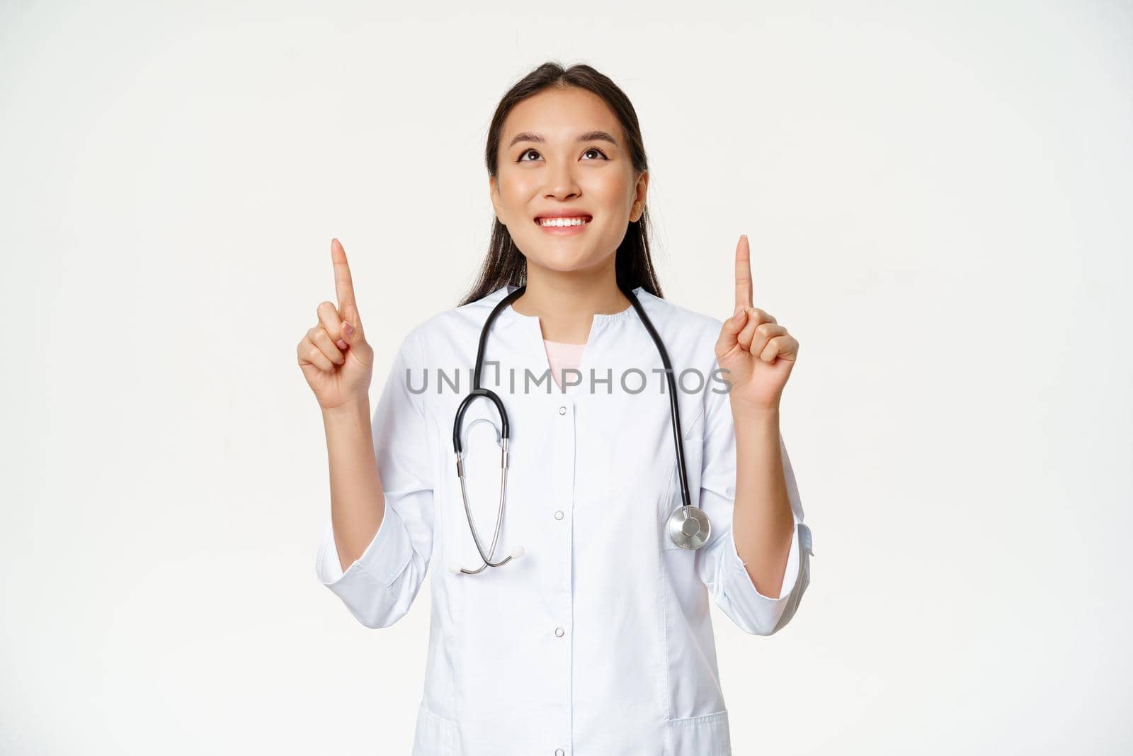 Happy smiling doctor, asian woman physician looking up with cheerful face expression, wearing medical uniform, white background.