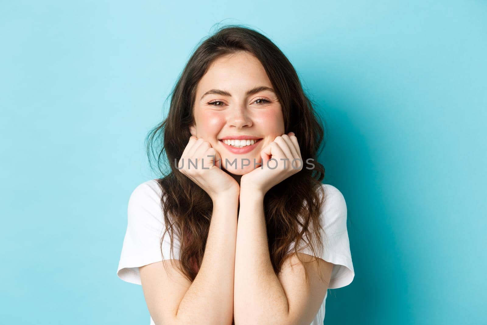 Close up portrait of cheerful woman smiling with teeth, lean face on hands and admiring something, looking with delight, standing happy against blue background.