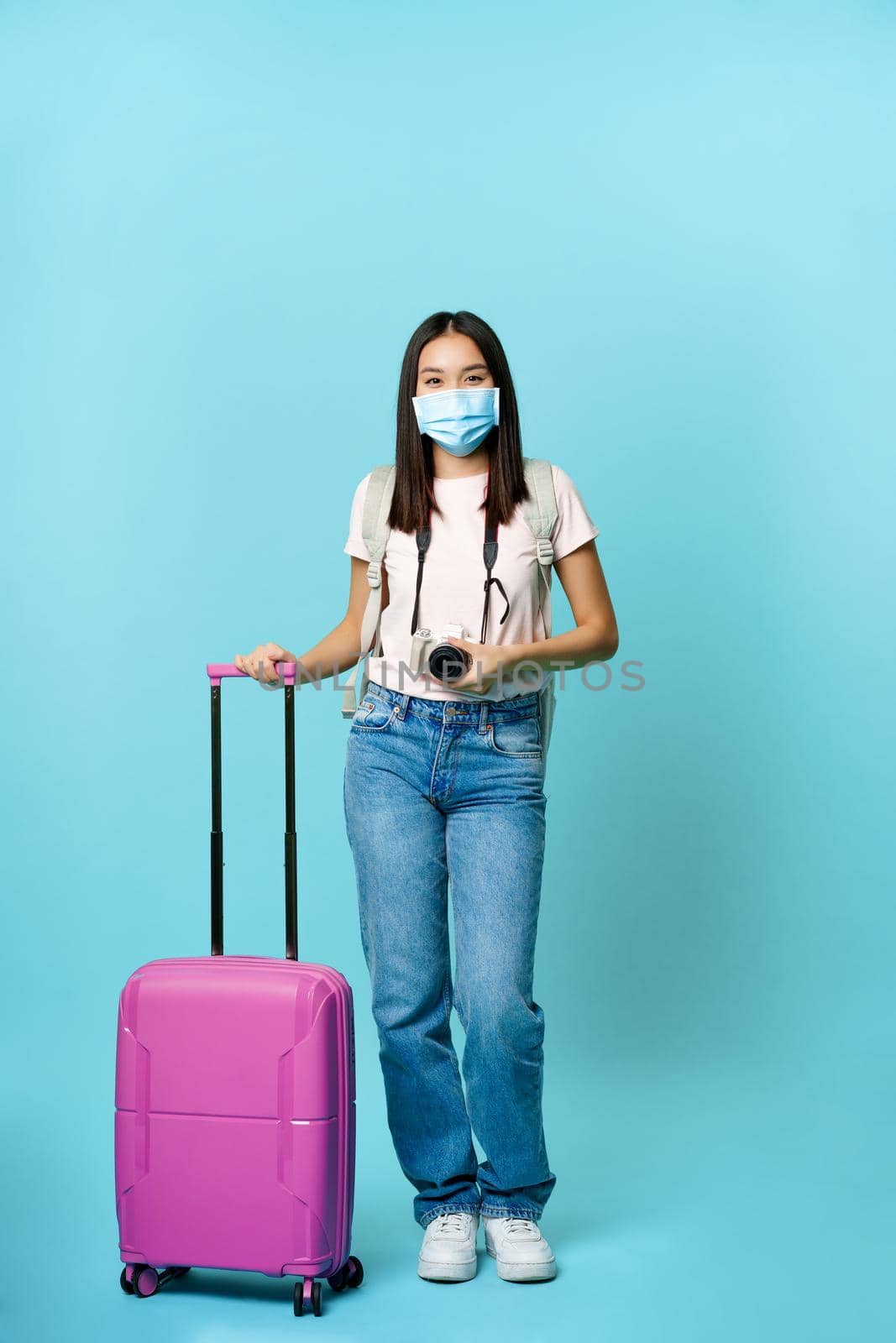 Happy asian girl traveller, tourist with suitcase and camera, going on vacation, wearing medical face mask during covid-19 restrictions, blue background by Benzoix
