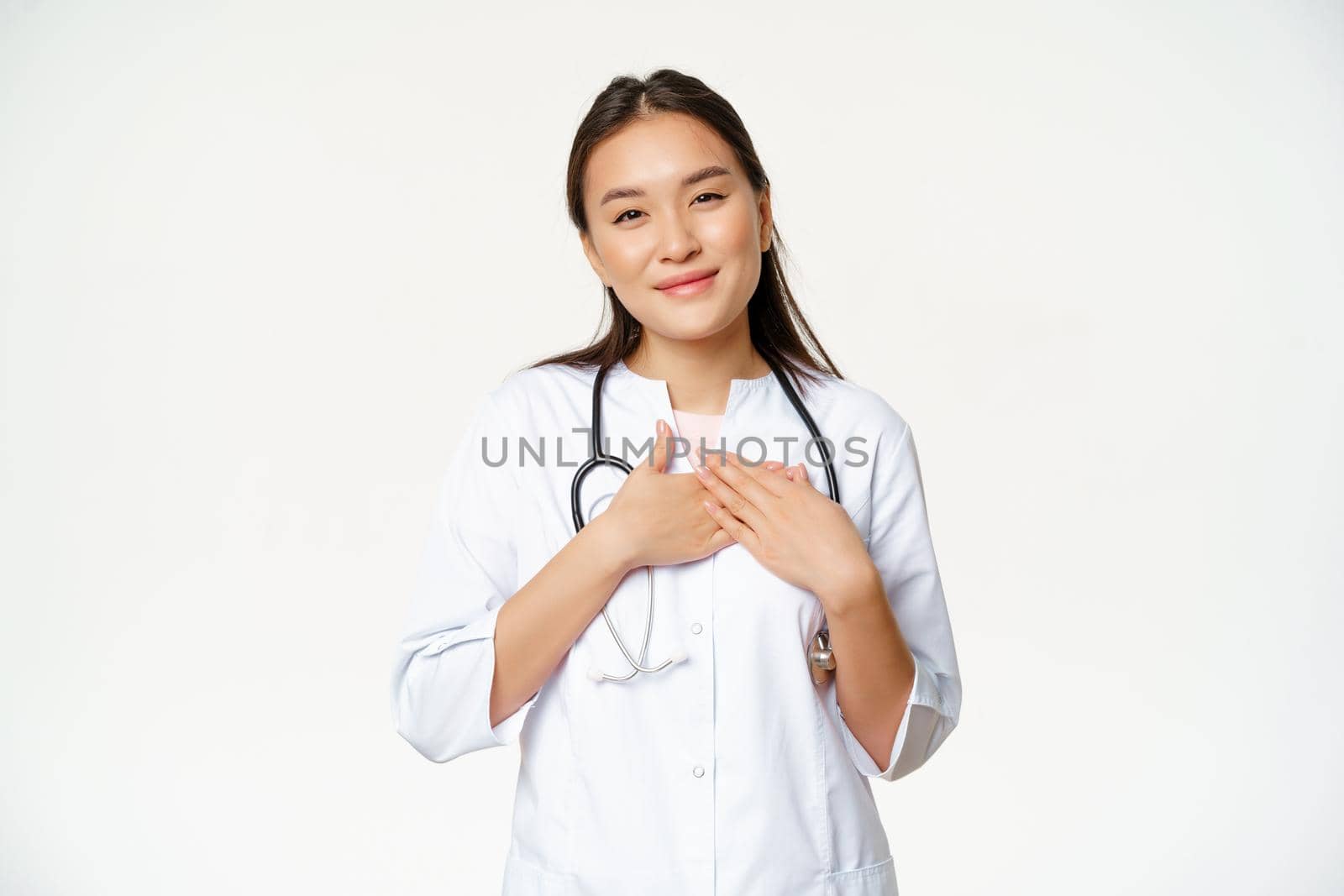 Image of female doctor caring for patients, holding hands on heart, smiling pleasant, standing in medical uniform against white background.