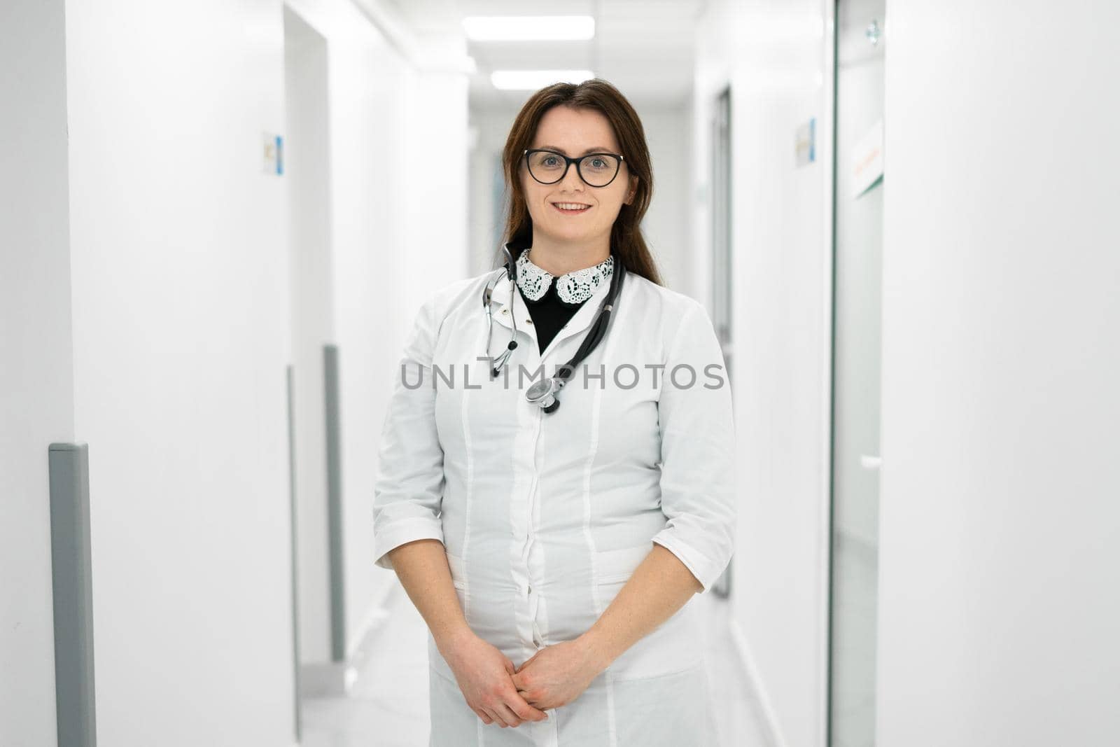 Headshot portrait of smiling millennial female doctor wearing medical uniform and stethoscope looking at camera in the corridor of a modern hospital. Healthcare concept, medical insurance by Tomashevska