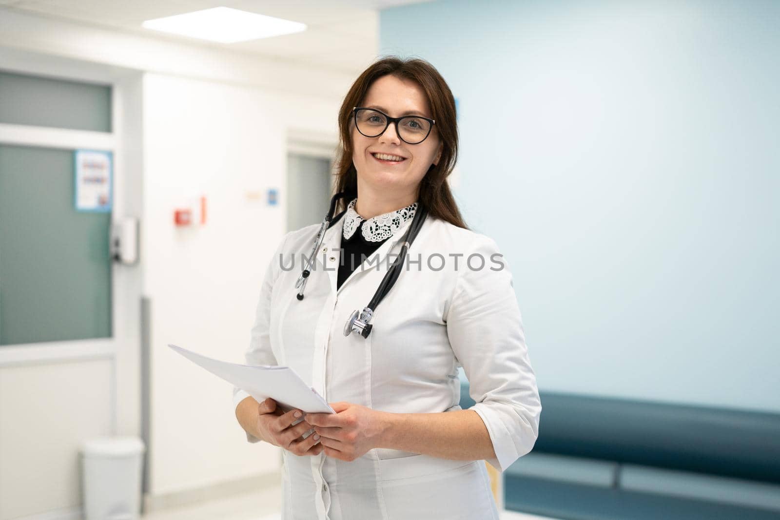 Headshot portrait of smiling millennial female doctor wearing medical uniform and stethoscope looking at camera in the corridor of a modern hospital. Healthcare concept, medical insurance.