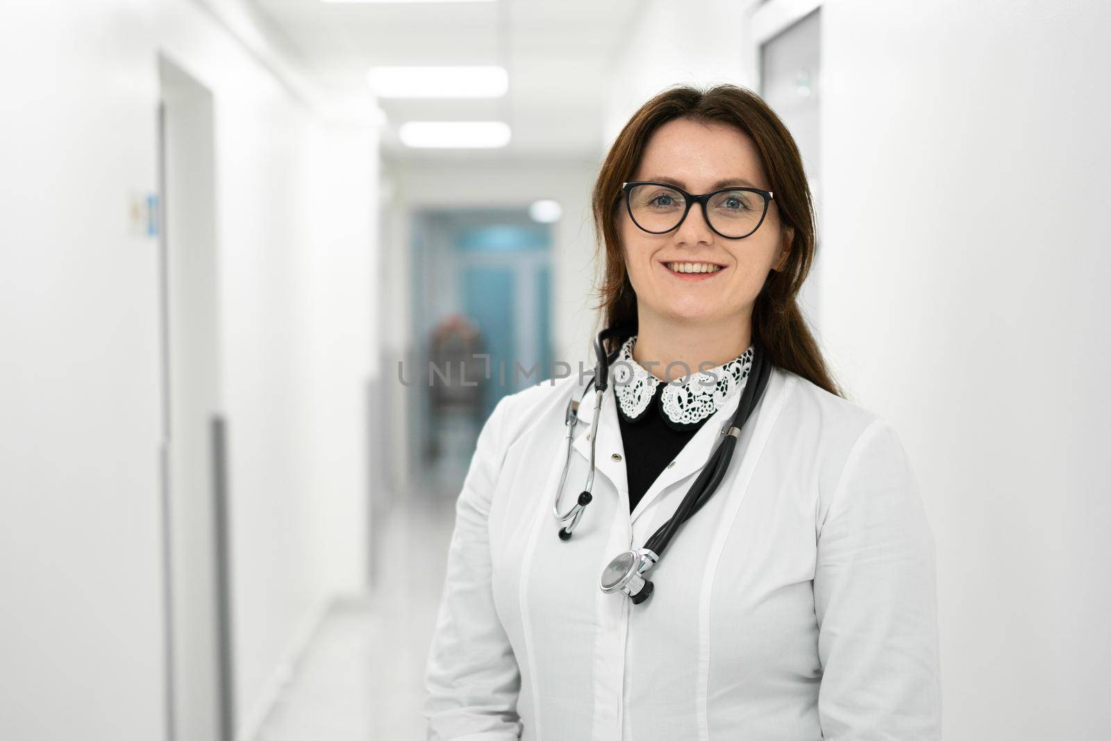 Headshot portrait of smiling millennial female doctor wearing medical uniform and stethoscope looking at camera in the corridor of a modern hospital. Healthcare concept, medical insurance by Tomashevska