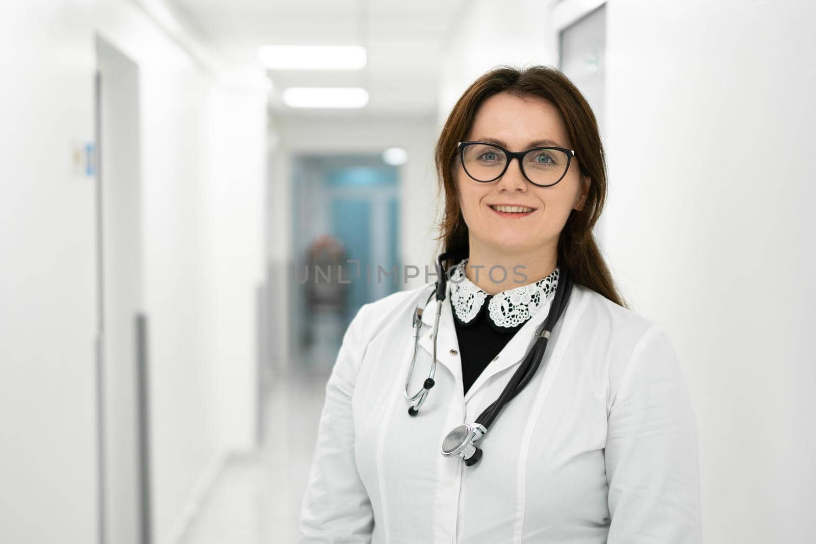 Female doctor standing in hospital corridor. Portrait of a young woman doctor in glasses and a white coat posing in a modern clinic. Proud professional woman doctor therapist looking at camera.