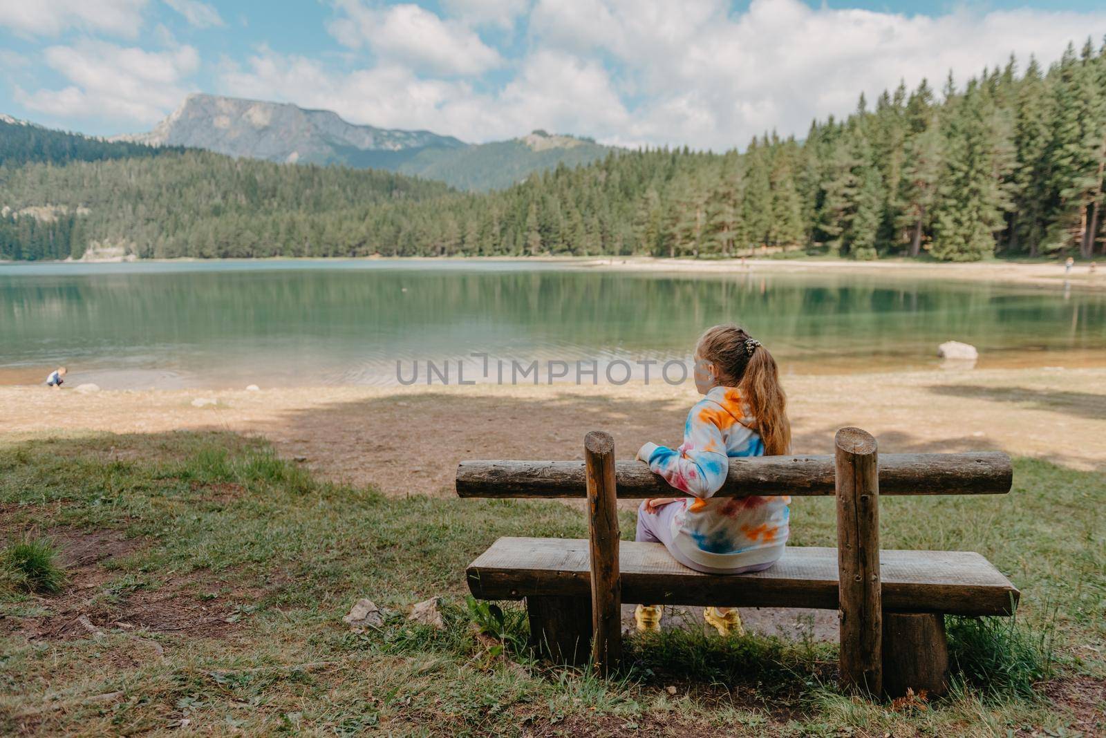 Young woman on shore of mountain lake. The back of girl against background of mountains. Young girl sitting near by beautiful Black Lake in Montenegro. Original wallpaper from summer morning.