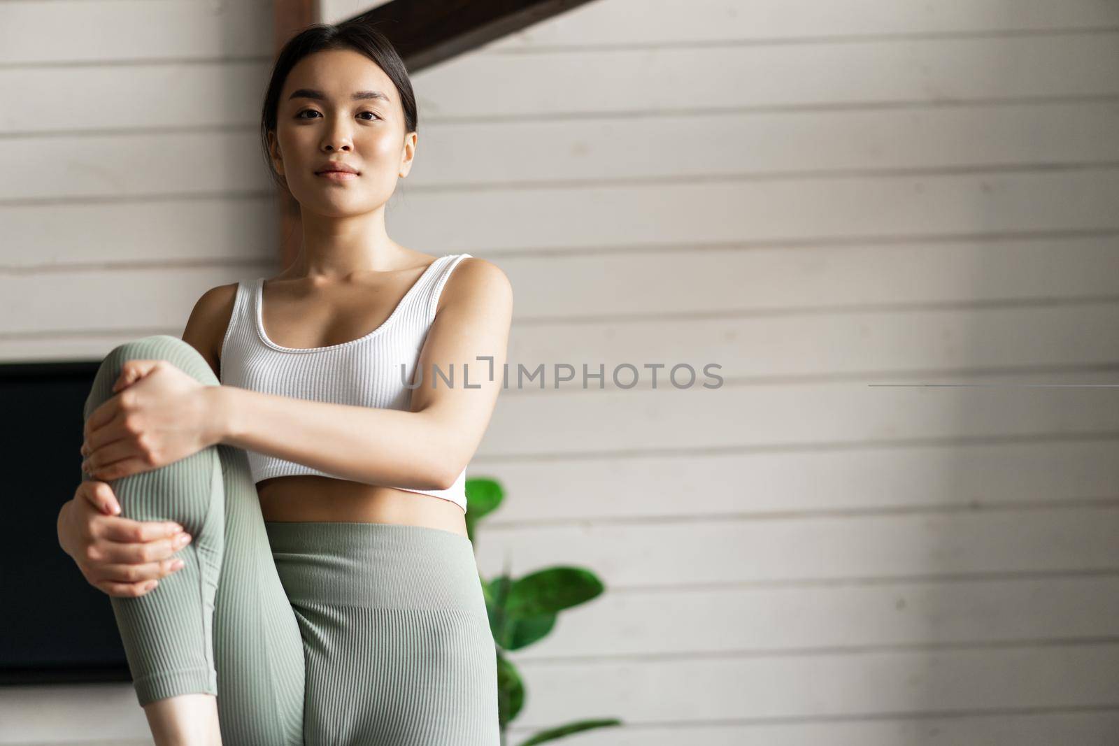 Portrait of asian girl doing workout at home, lifting legs and looking at distance, exercising in living room without fitness gear.