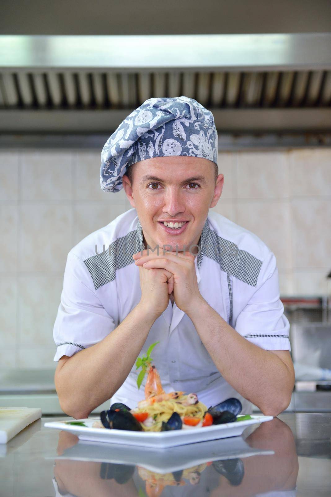 Handsome chef dressed in white uniform decorating pasta salad and seafood fish in modern kitchen