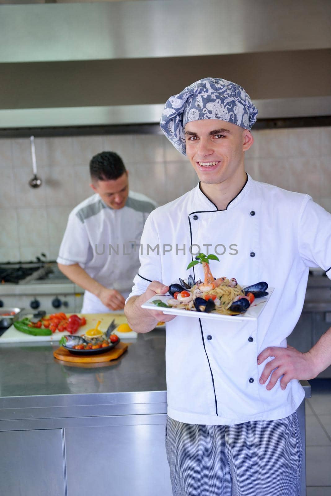 Handsome chef dressed in white uniform decorating pasta salad and seafood fish in modern kitchen