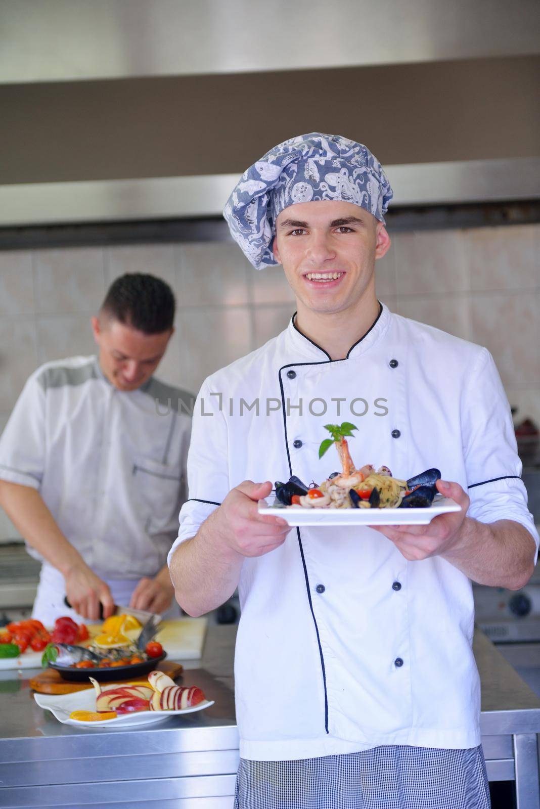 Handsome chef dressed in white uniform decorating pasta salad and seafood fish in modern kitchen