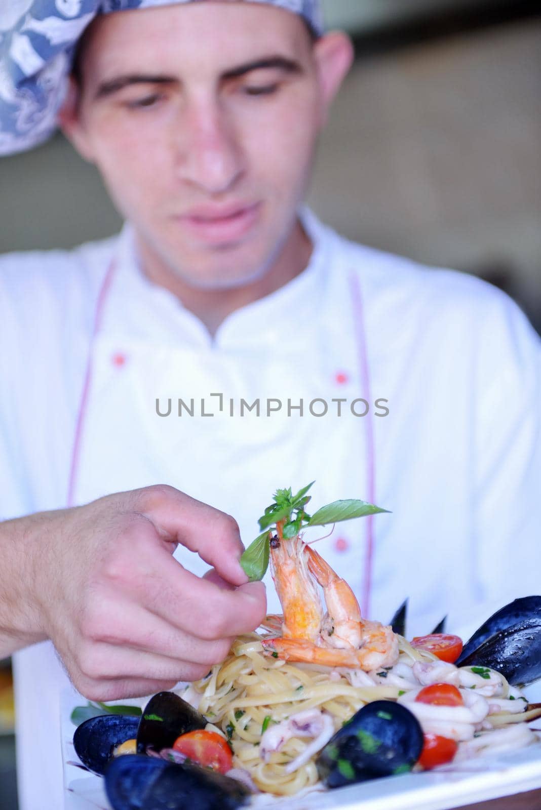 Handsome chef dressed in white uniform decorating pasta salad and seafood fish in modern kitchen