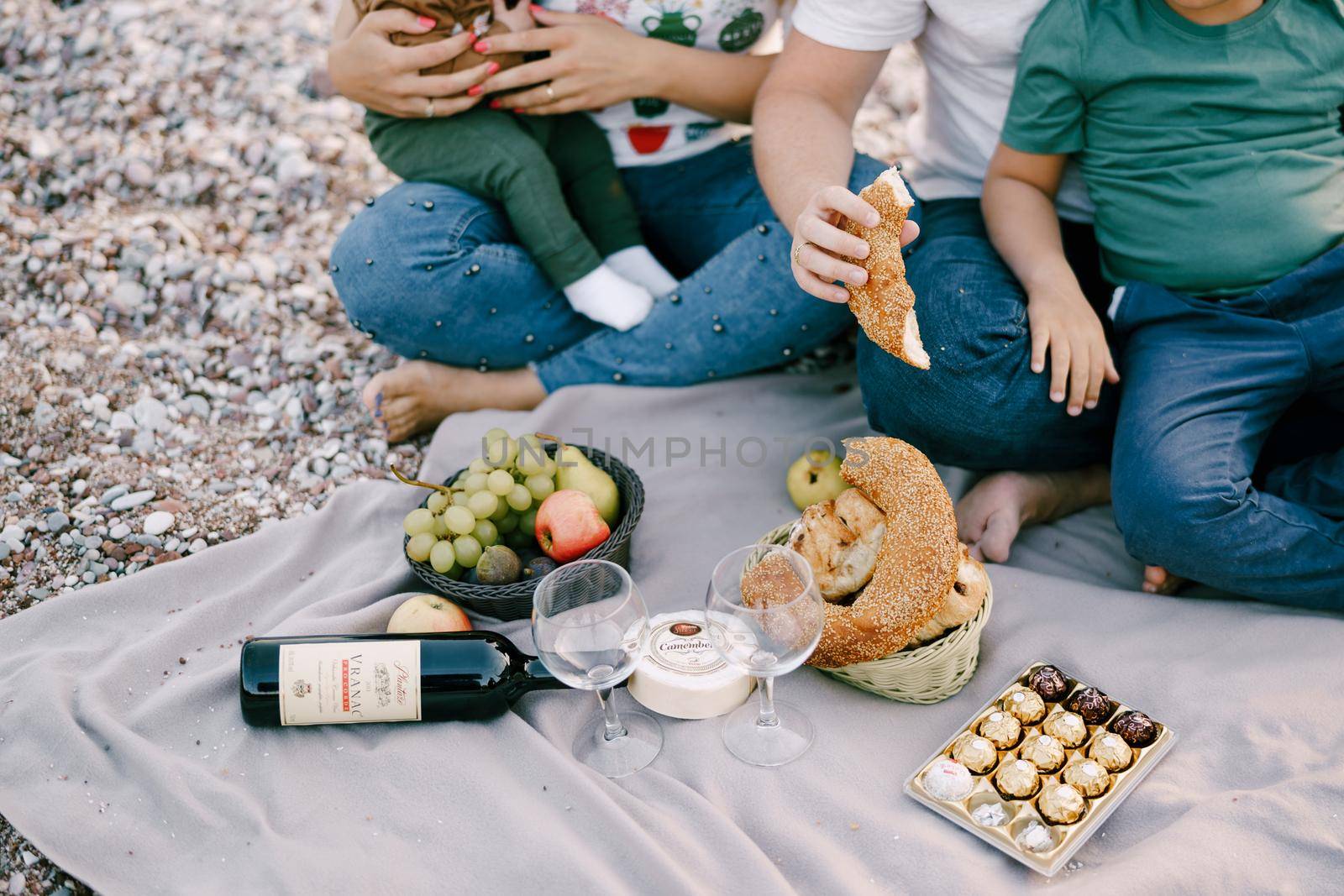 Man and woman with children on the beach sit on a blanket with food by Nadtochiy