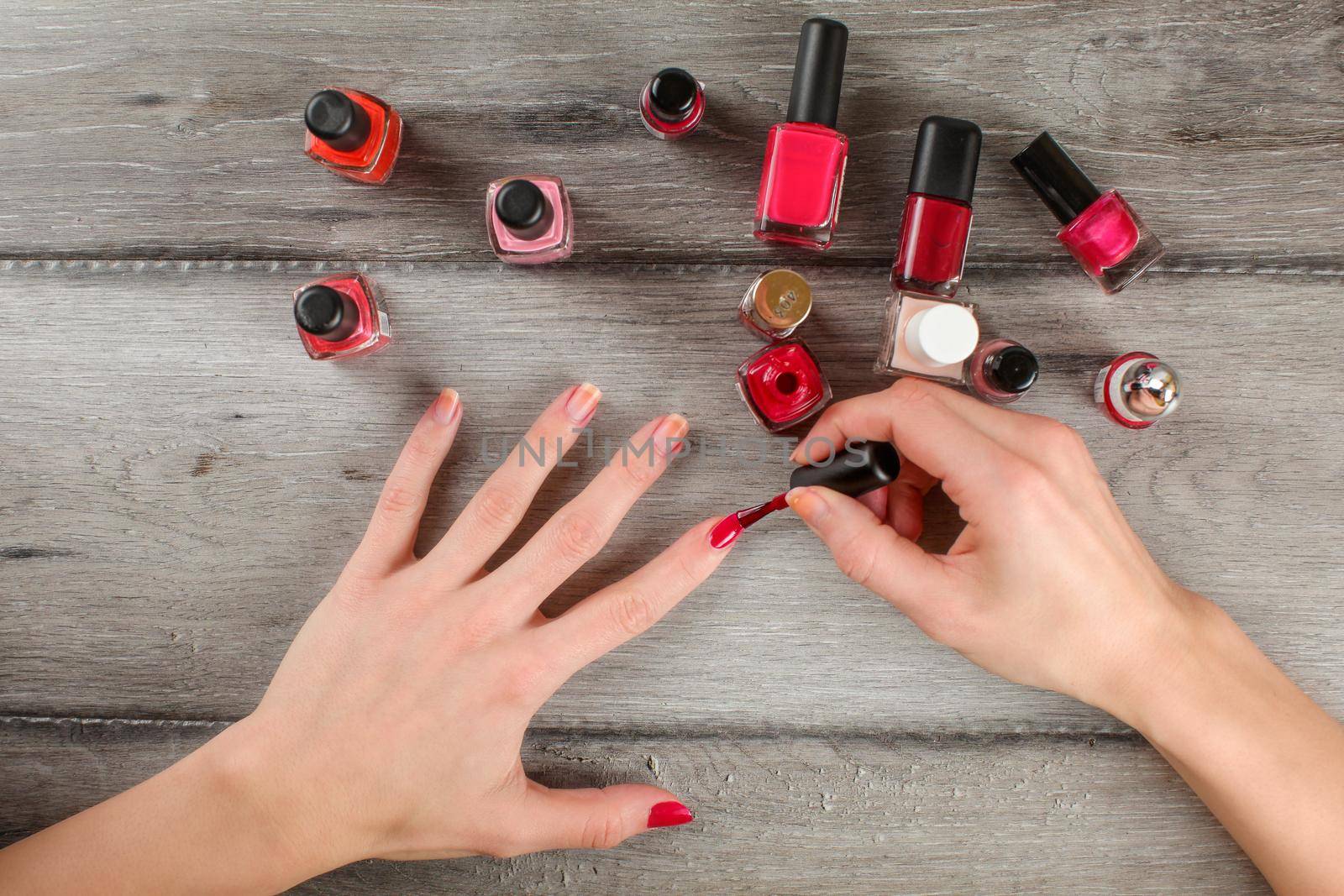 Top view to woman hands, applying coat of red nail varnish on her nails, with lot of nail polish on gray wood desk in background.