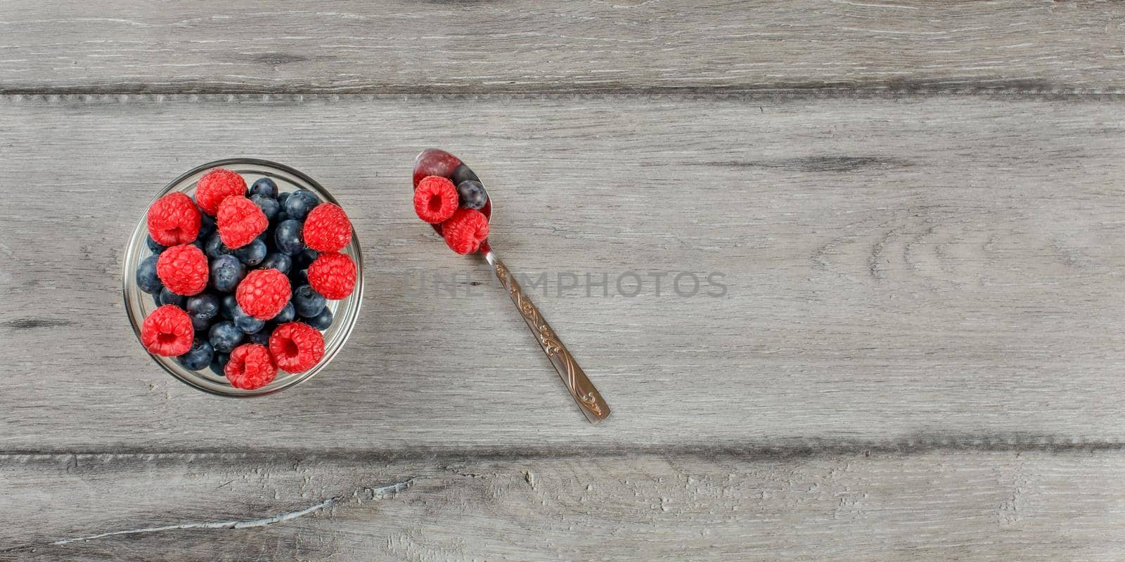 Table top view on small glass bowl filled with mix of blueberries and raspberries, silver spoon next to it, suggesting healthy snack is ready to eat. by Ivanko