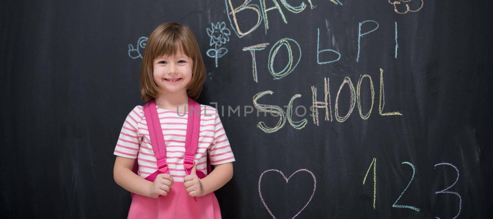 Happy school girl child with backpack writing  back to school on black chalkboard