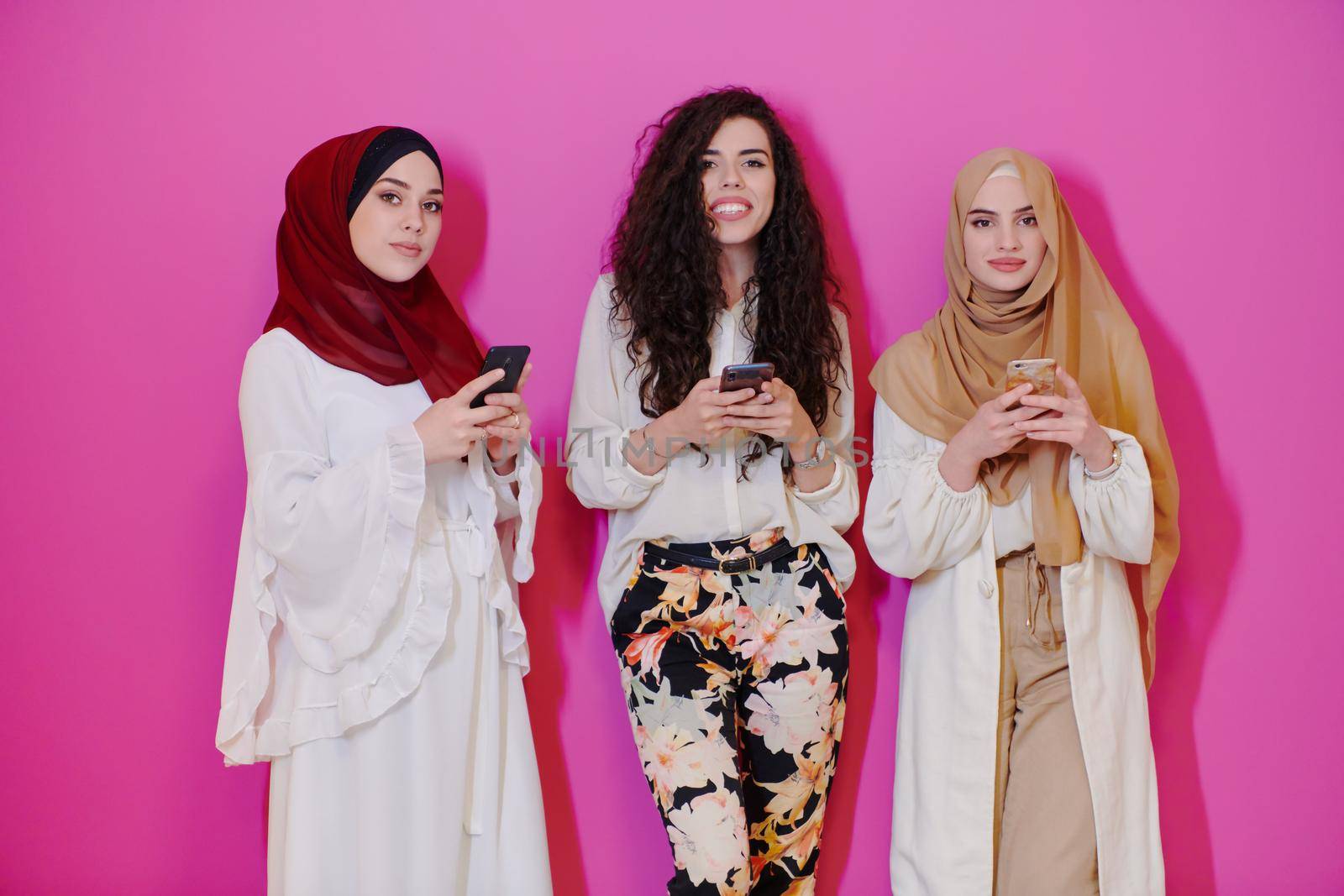 group of beautiful muslim women two of them in fashionable dress with hijab using mobile phone isolated on pink background representing modern islam fashion technology and ramadan kareem concept