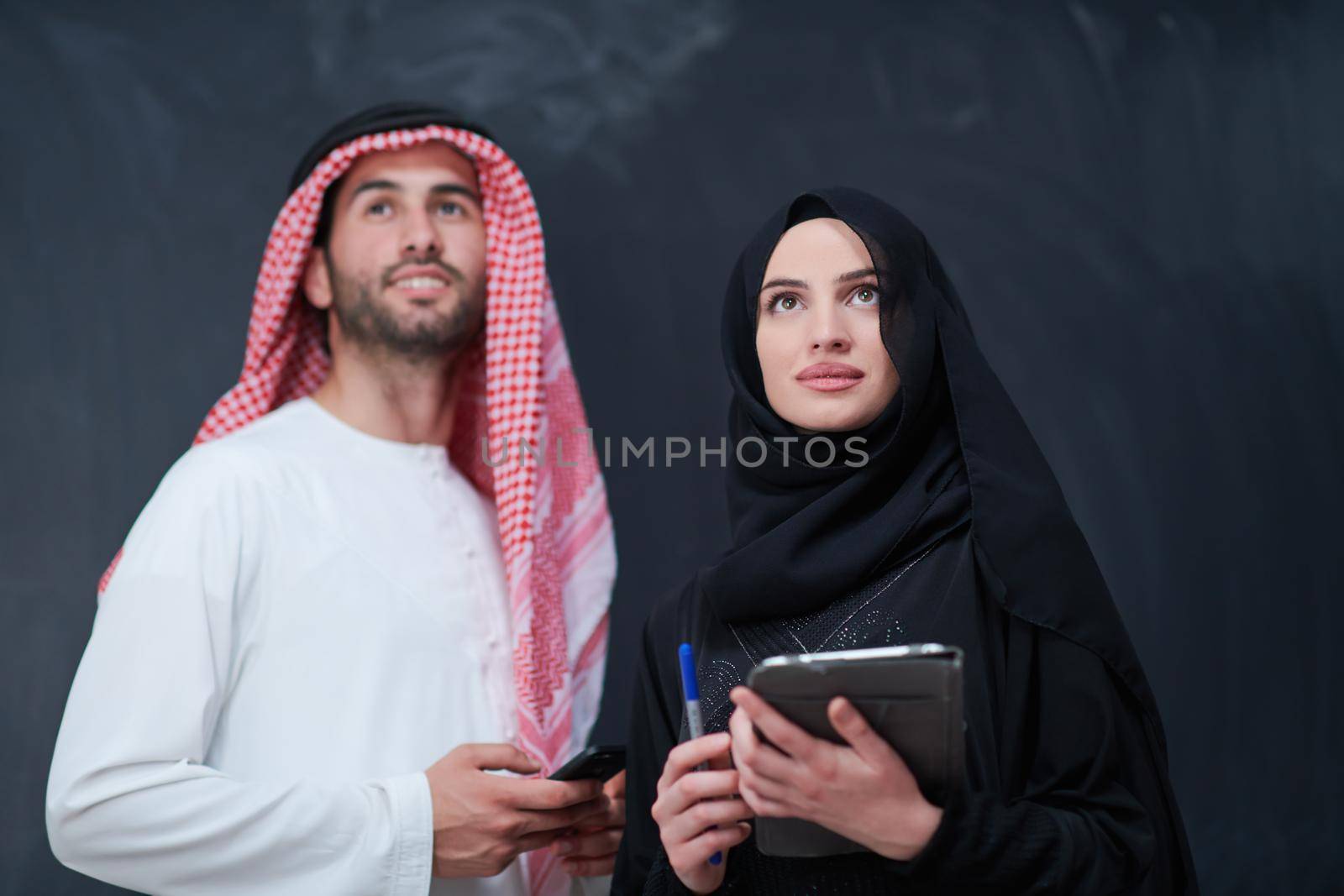 young muslim business couple arabian man with woman in fashionable hijab dress using mobile phone and tablet computer in front of black chalkboard representing modern islam fashion technology and ramadan kareem concept