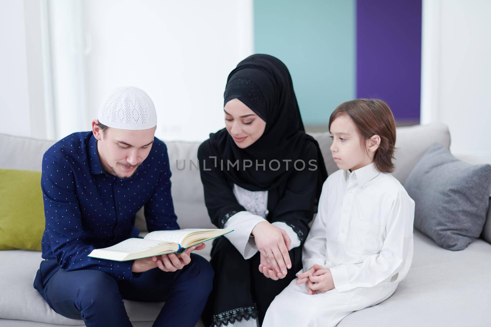 Traditional muslim family parents with children reading Quran and praying together on the sofa before iftar dinner during a ramadan feast at home