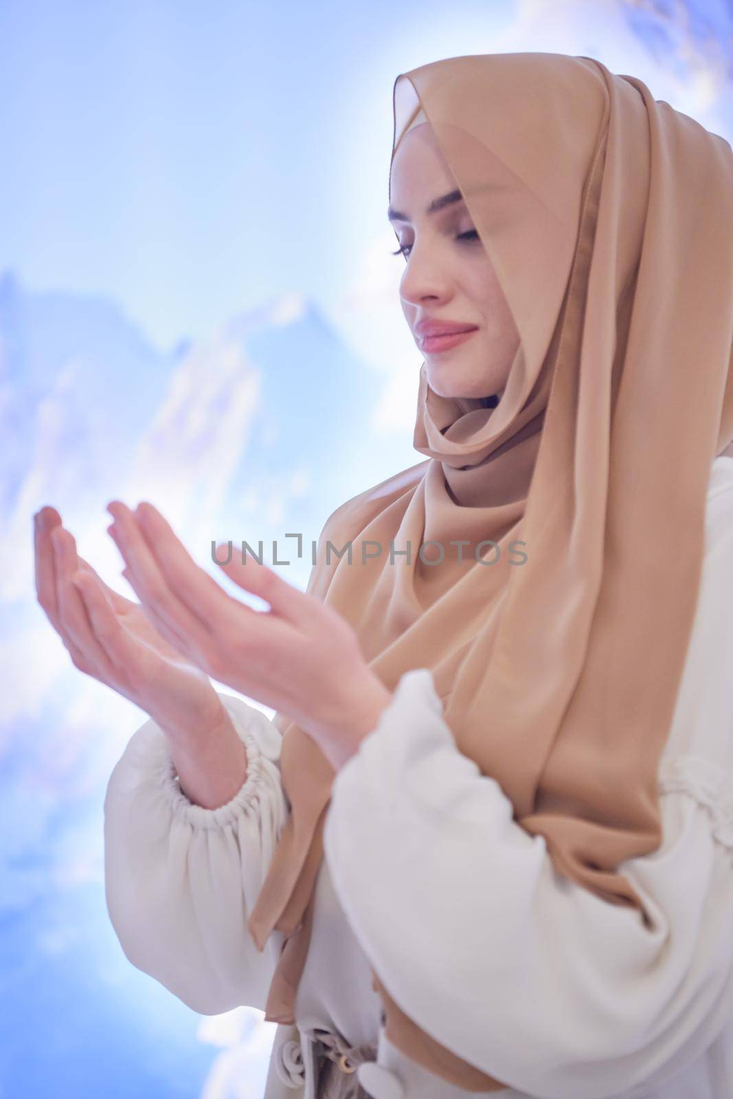 portrait of beautiful muslim woman in fashionable dress with hijab making traditional prayer to God, keeps hands in praying gesture isolated on modern winter mountains background representing modern islam fashion and ramadan kareem concept