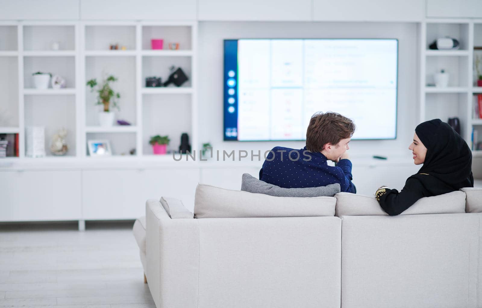 young muslim couple woman wearing islamic hijab clothes sitting on sofa watching TV together during the month of Ramadan at modern home