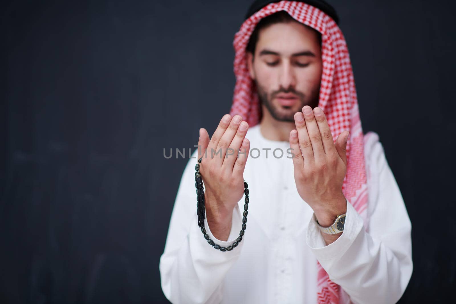 young arabian man in traditional clothes making traditional prayer to God, keeps hands in praying gesture in front of black chalkboard representing modern islam fashion and ramadan kareem concept