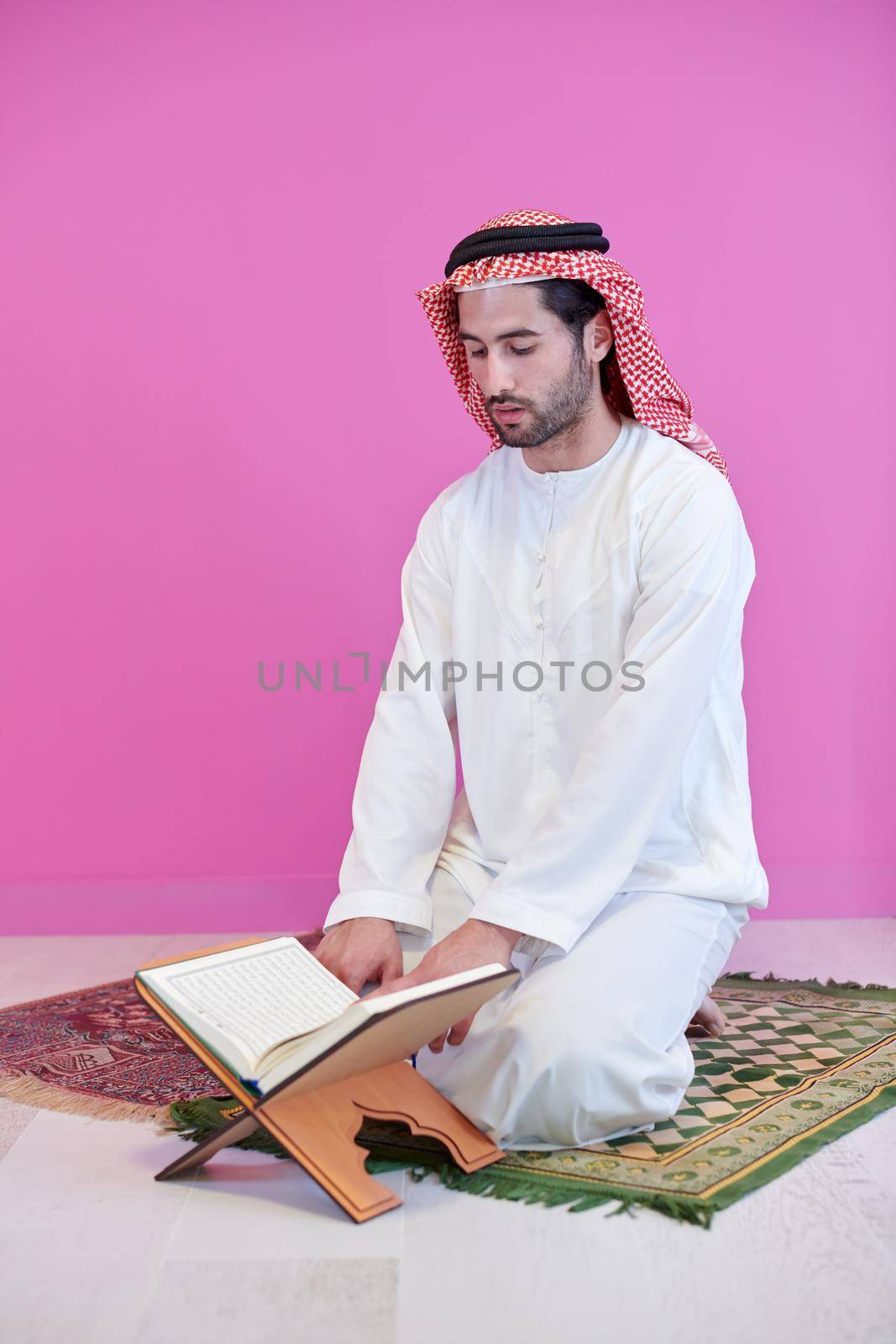 young arabian muslim man in traditional clothes reading holy book Quran on the praying carpet in front of pink wall before iftar dinner during a ramadan feast at home