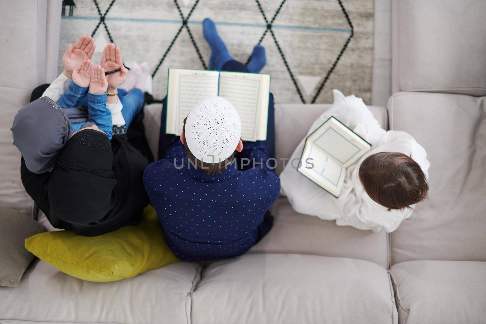 Traditional muslim family parents with children reading Quran and praying together on the sofa before iftar dinner during a ramadan feast at home top view