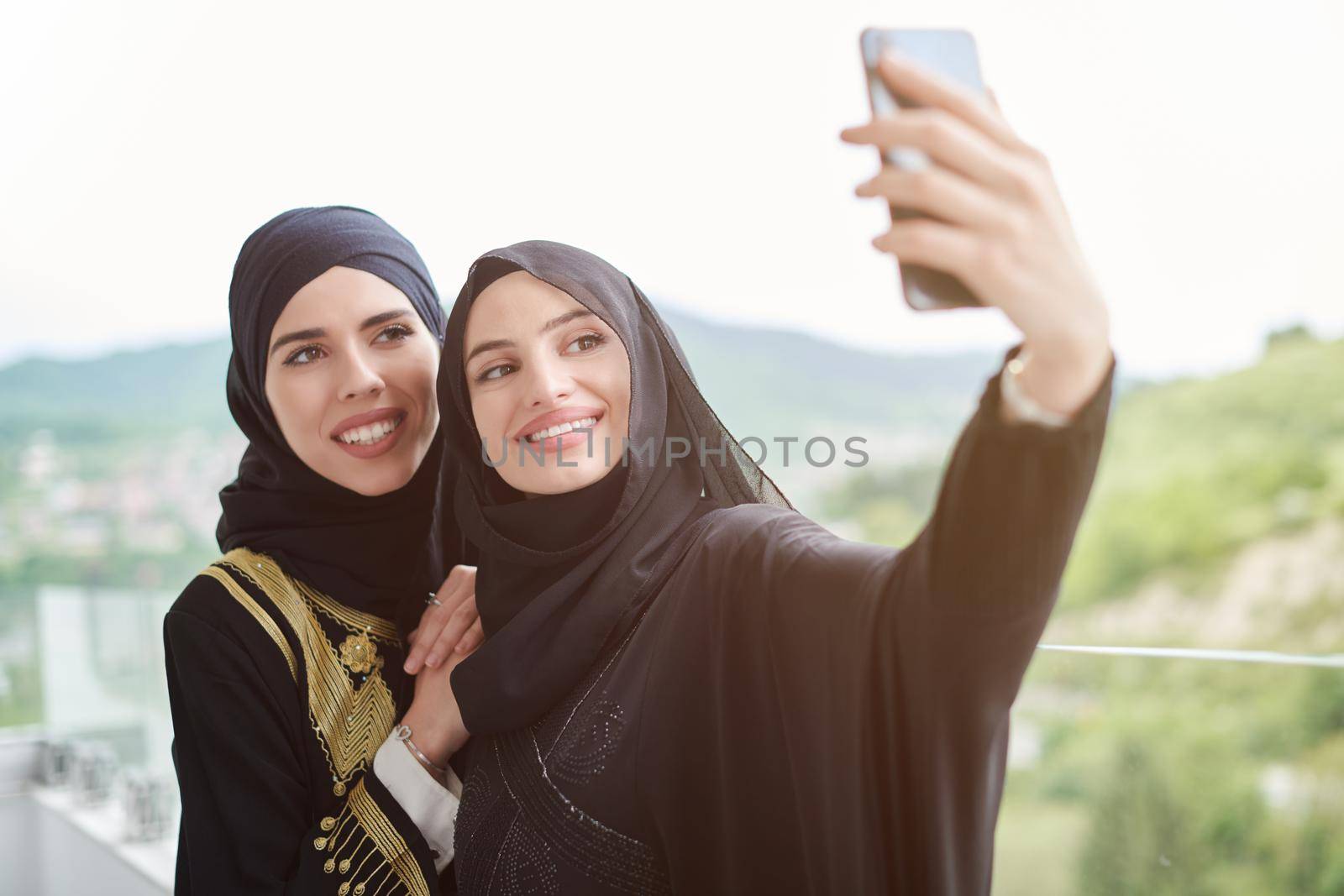 young beautiful muslim women in fashionable dress with hijab using mobile phone while taking selfie picture on the balcony representing modern islam fashion technology and ramadan kareem concept