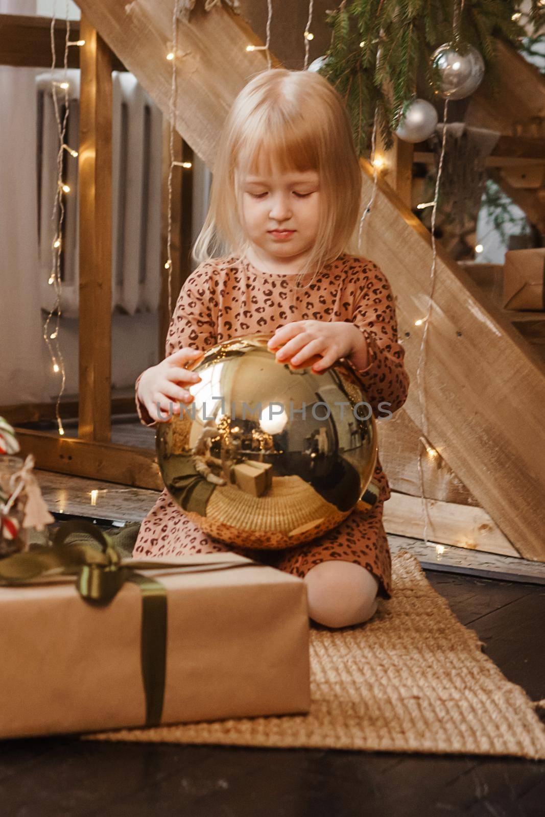 A little blonde girl is sitting next to a wooden staircase in a Scandinavian interior decorated in a New Year's style. A child holds a golden Christmas ball.