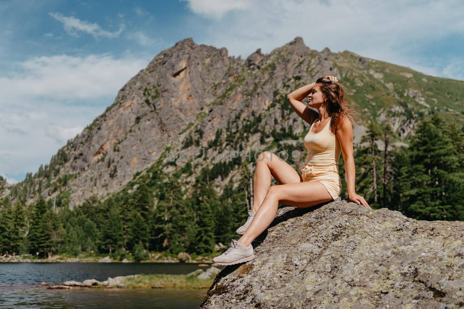 Tourist girl enjoys the magical view of the lake, coniferous forest and magical view sitting on big stone on the shore of a turquoise lake in the mountains. Hiking in the Natural Park. Cute girl tourist sitting on a large stone by the lake. The water is clear, stones are visible under the water. by Andrii_Ko