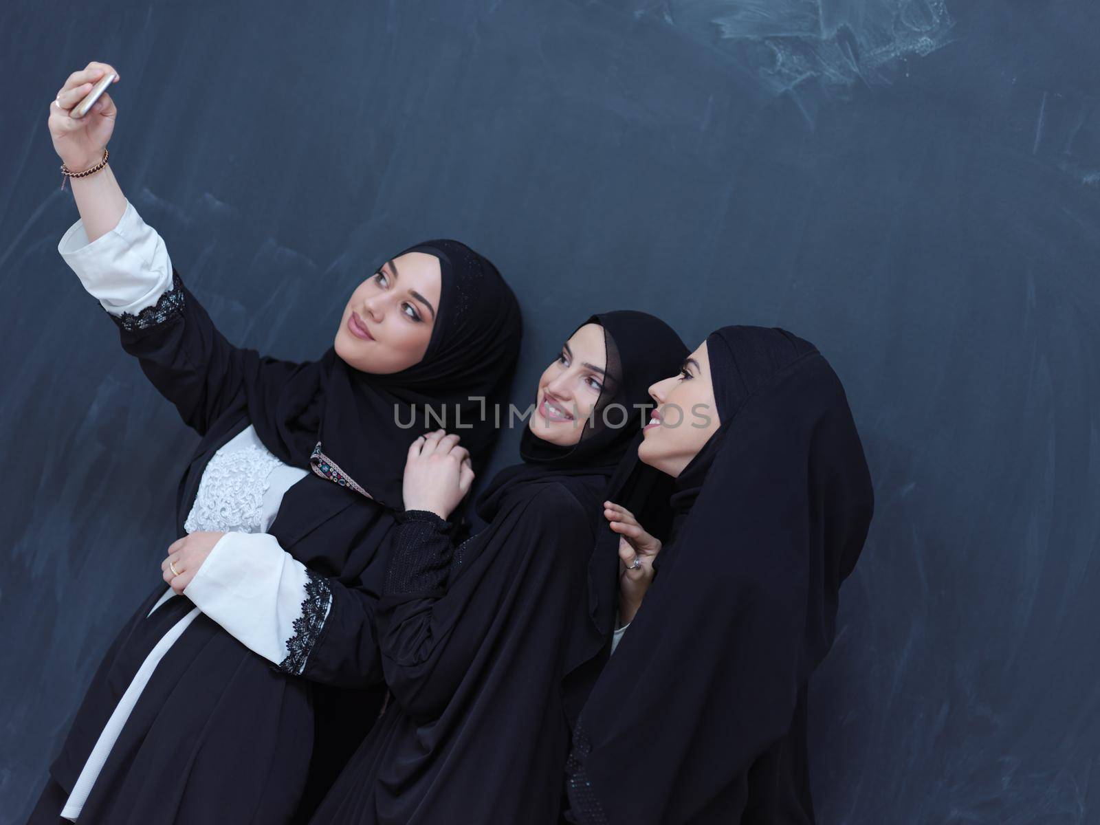 group of young beautiful muslim women in fashionable dress with hijab using mobile phone while taking selfie picture in front of black chalkboard representing modern islam fashion technology and ramadan kareem concept