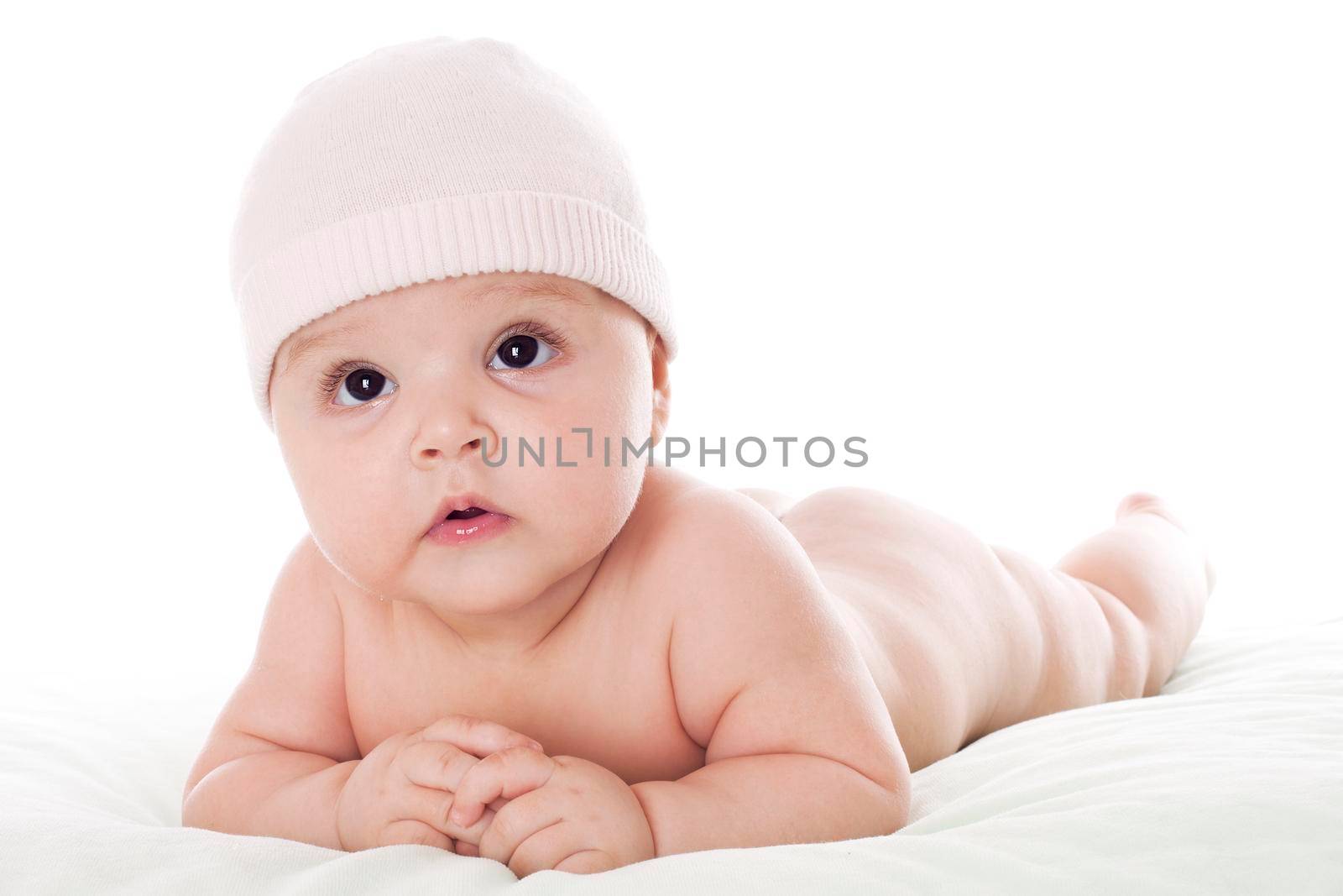 Cute baby lying on stomach on white floor background, wearing a cap and looking up being greatly surprising.