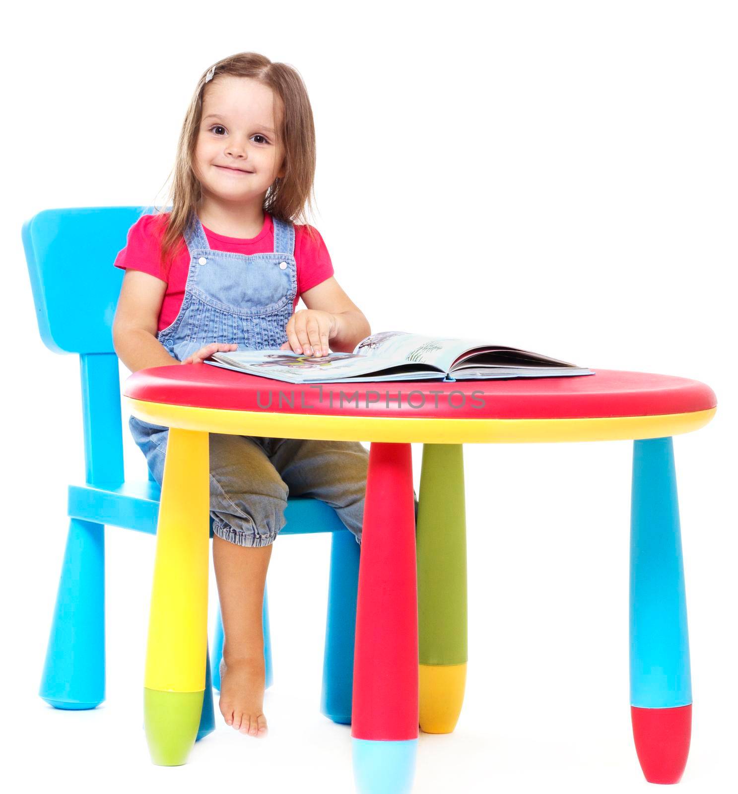 three years old girl child sitting at the table and reading a book, isolated on white