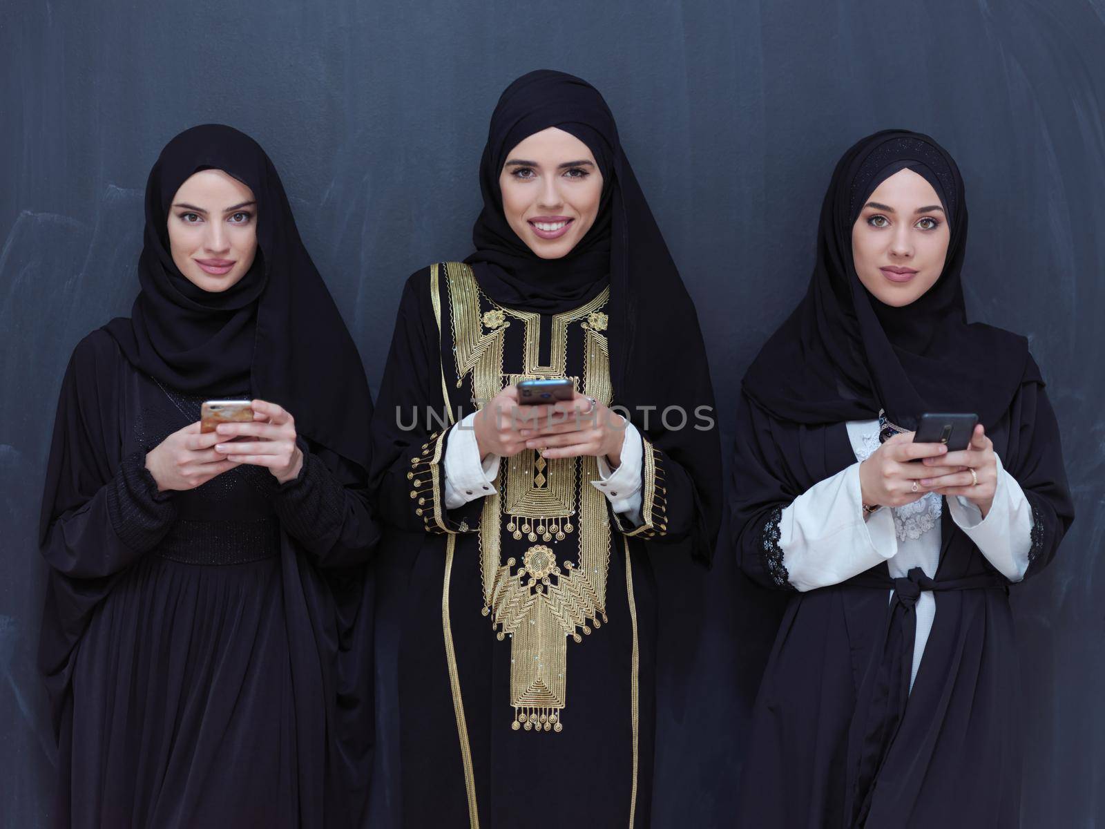 group of beautiful muslim women in fashionable dress with hijab using mobile phone in front of black chalkboard representing modern islam fashion technology and ramadan kareem concept