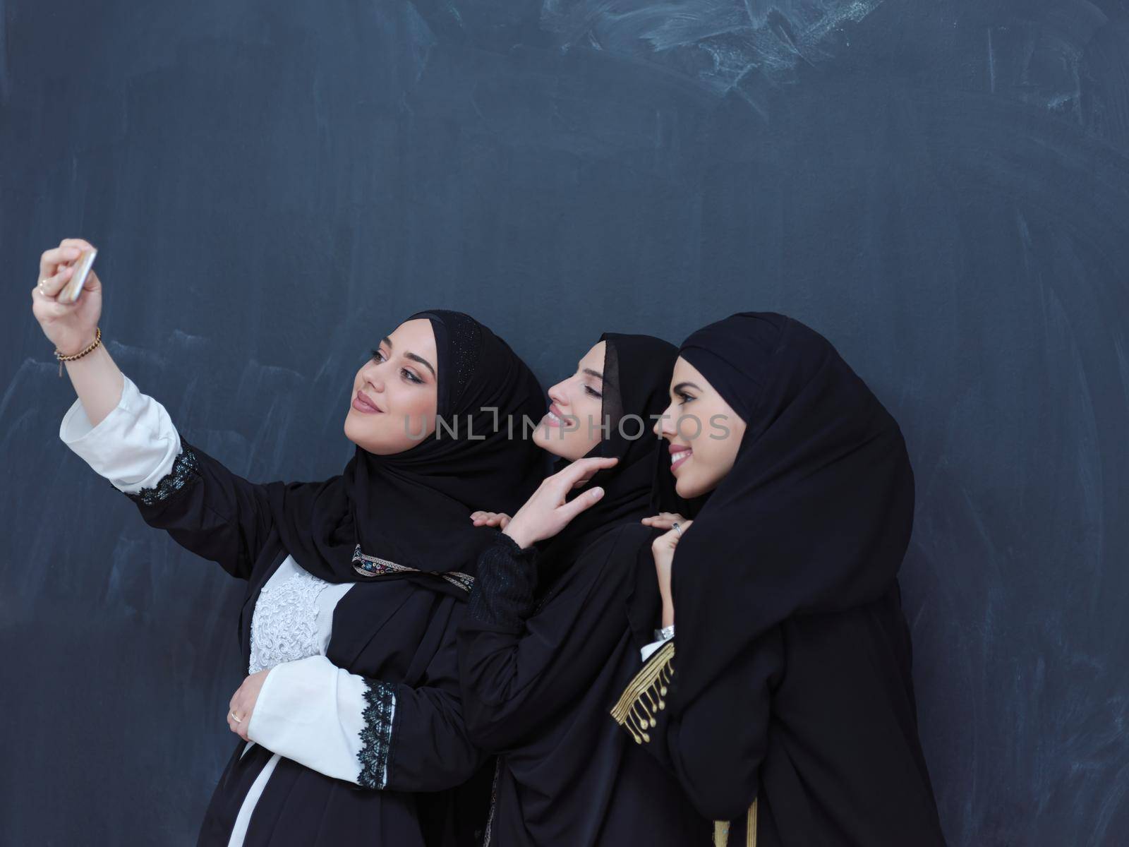 group of young beautiful muslim women in fashionable dress with hijab using mobile phone while taking selfie picture in front of black chalkboard representing modern islam fashion technology and ramadan kareem concept