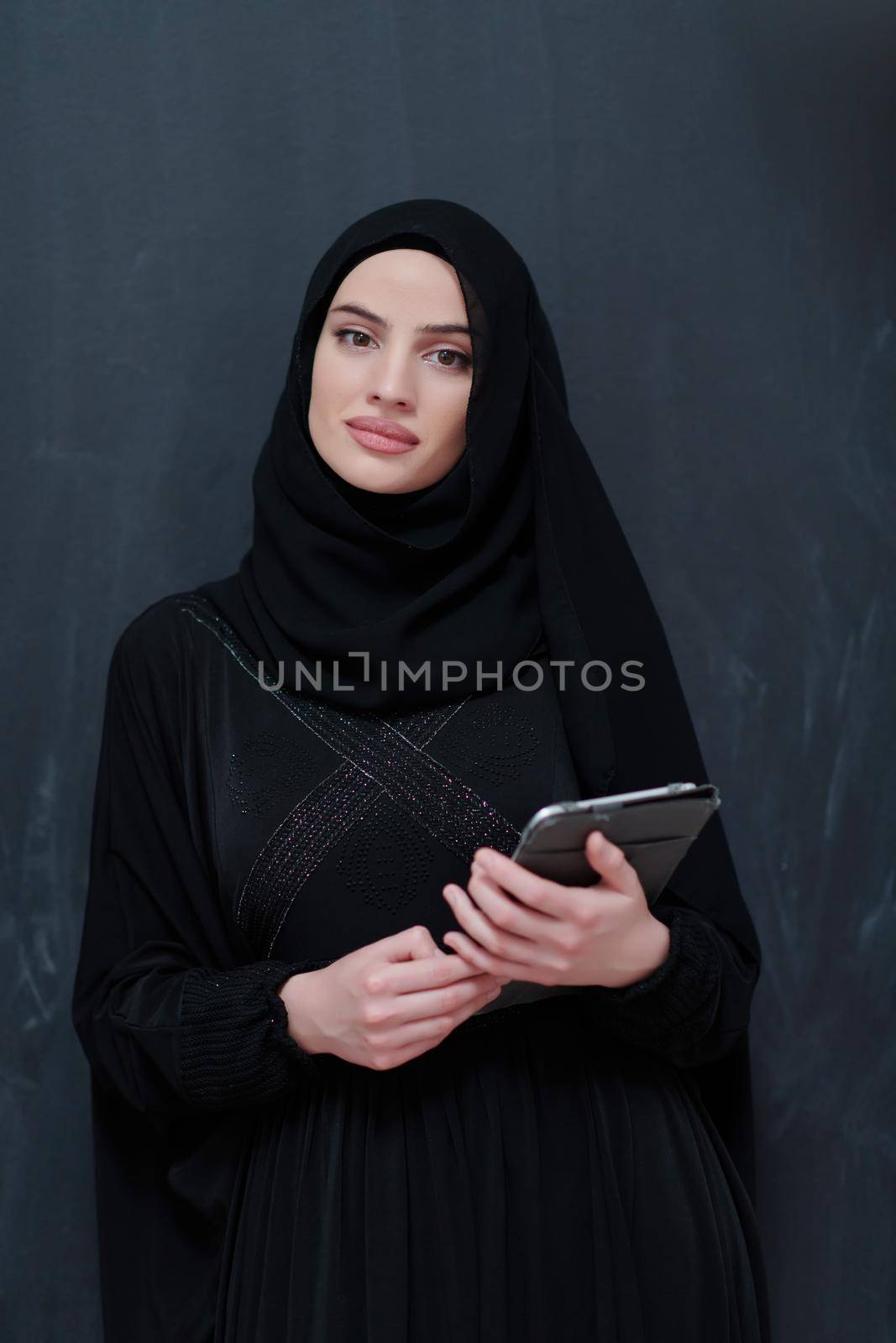 Young Arab businesswoman in traditional clothes or abaya with tablet computer in front of black chalkboard representing modern islam fashion technology